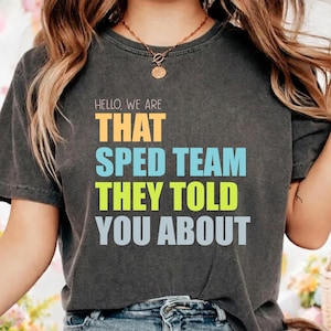 Puede incluir: Camiseta gris oscuro con el texto "HELLO, WE ARE THAT SPED TEAM THEY TOLD YOU ABOUT" en naranja, azul, verde y gris. La camiseta tiene cuello redondo y mangas cortas.