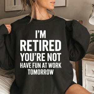 Może przedstawiać: Czarna bluza z białym napisem "I'M RETIRED YOU'RE NOT HAVE FUN AT WORK TOMORROW". Bluza ma okrągły dekolt i długie rękawy. Zdjęcie zrobione w pomieszczeniu.