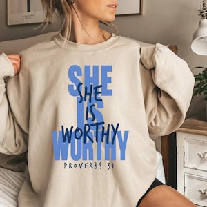 May include: Beige sweatshirt with the text "SHE IS WORTHY" in blue block letters. The words "PROVERBS 31" are printed below the main text. The sweatshirt is a neutral color and has long sleeves.