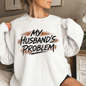 May include: White sweatshirt featuring the text "MY HUSBAND'S PROBLEM" in black, with a brown brushstroke background. A casual, comfortable style of clothing.