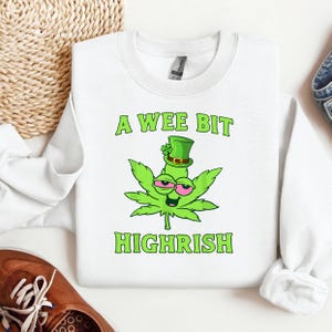 Può includere: Felpa bianca con la scritta "A Wee Bit Highrish" in verde. Il design presenta una foglia di cannabis verde con un cappello da folletto e occhi rosa. La felpa è del marchio Gildan.