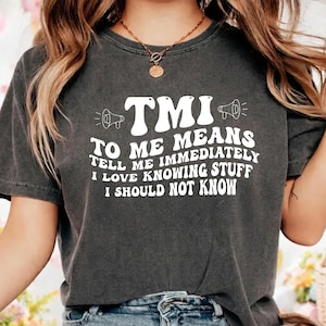 May include: Dark gray t-shirt with the white text "TMI TO ME MEANS TELL ME IMMEDIATELY I LOVE KNOWING STUFF I SHOULD NOT KNOW." The shirt has a relaxed fit and short sleeves.
