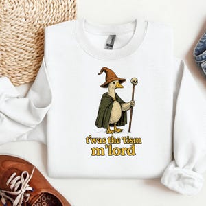 Op de afbeelding: Wit sweatshirt met een cartoon eend verkleed als tovenaar, bruine hoed en groene cape, met een staf met een schedel. De tekst "t'was the 'tism m'lord" staat eronder.