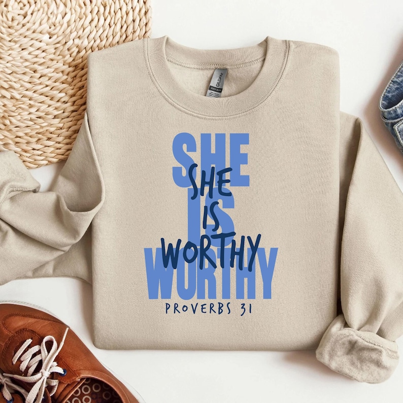 May include: A beige crewneck sweatshirt with the words "SHE IS WORTHY" in blue block letters. The text "PROVERBS 31" is printed below the main text. The sweatshirt is folded on a light surface.