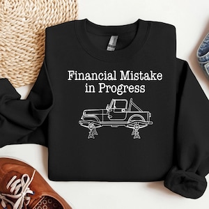 Op de afbeelding: Zwart sweatshirt met de tekst "Financial Mistake in Progress" boven een witte lijntekening van een Jeep op krikken. Het sweatshirt ligt op een lichtgekleurd oppervlak, met een bruine schoen en denim jeans zichtbaar.