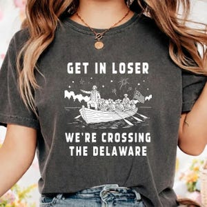 May include: Dark gray t-shirt with the text "GET IN LOSER WE'RE CROSSING THE DELAWARE" in white. The graphic depicts a boat with people and an American flag. The shirt has a vintage, worn look.