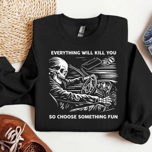 以下が含まれることがあります： 車を運転する骸骨の白いグラフィックが特徴の黒いスウェットシャツ。「EVERYTHING WILL KILL YOU SO CHOOSE SOMETHING FUN」の文字がグラフィックの上と下にプリントされています。スウェットシャツは折りたたまれています。