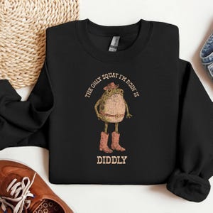 May include: Black sweatshirt featuring a cartoon frog wearing a cowboy hat, belt, and boots. The text reads "THE ONLY SQUAT I'M DOIN IS DIDDLY" in a circular and stacked layout. The frog is tan and the boots are a reddish-brown color.