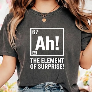 May include: Dark grey t-shirt featuring a white graphic with the text "67 Ah! 67" in a square, and "THE ELEMENT OF SURPRISE!" underneath. The shirt has a crew neck and short sleeves.