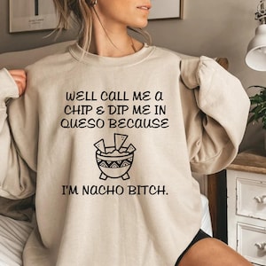 May include: Beige sweatshirt with black text that reads "WELL CALL ME A CHIP & DIP ME IN QUESO BECAUSE I'M NACHO BITCH." A graphic of a bowl of chips and dip is below the text. The sweatshirt is a casual style.