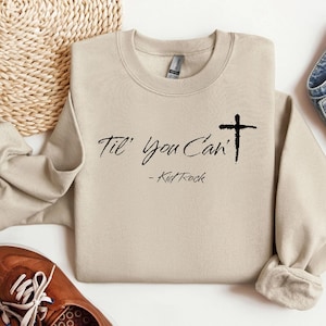 Puede incluir: Sudadera beige de cuello redondo con la frase "Til' You Can't" y una cruz negra. El texto "- Kid Rock" está debajo. La sudadera está doblada y tiene un estilo casual.