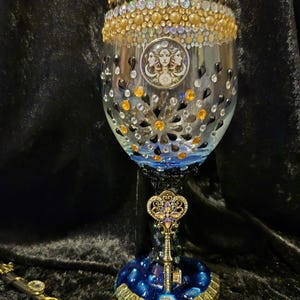 Puede incluir: Una copa de vino adornada y adornada con un tema celestial. La copa presenta un diseño detallado con cristales dorados, negros y transparentes, y un medallón central con una figura de tres caras. La base es azul oscuro con un adorno en forma de llave.