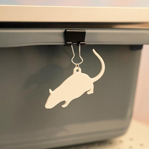 Könnte beinhalten: Ein weißer, rattenförmiger Anhänger hängt an einer schwarzen Büroklammer, die an einem grauen Behälter befestigt ist. Der Anhänger ist eine einfache Silhouette mit einem gebogenen Schwanz. Der Hintergrund ist eine weiche Mischung aus Rosa und Blau.