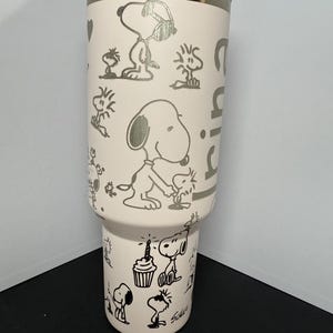 May include: A light pink tumbler featuring Snoopy and other Peanuts characters in silver etched designs. The tumbler has a silver rim and a clear lid. The design includes Snoopy with a birthday cake and other celebratory images.