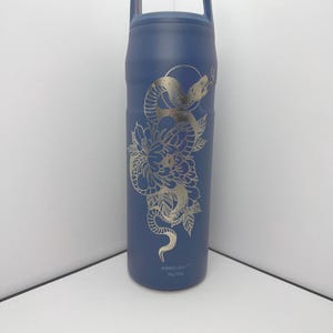 May include: A navy blue insulated water bottle with a silver snake and floral design. The bottle has a handle and the text "ABROUGHT" near the bottom. The design features a snake coiled around a flower.