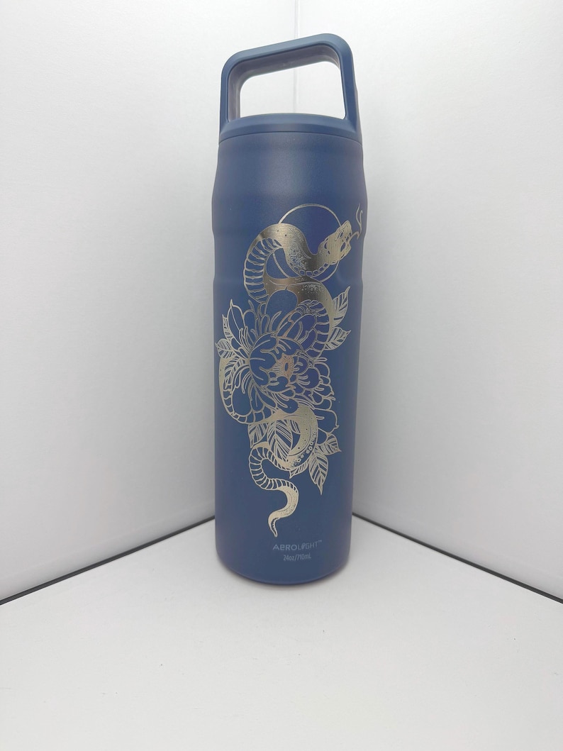 May include: A navy blue insulated water bottle with a silver snake and floral design. The bottle has a handle and the text "ABROUGHT" near the bottom. The design features a snake coiled around a flower.