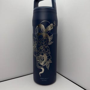 May include: A navy blue insulated water bottle with a silver floral and snake design. The bottle has a handle and the text "ABROLIGHT" and "740/750ml" printed on the bottom. The design features a snake coiled around a flower.