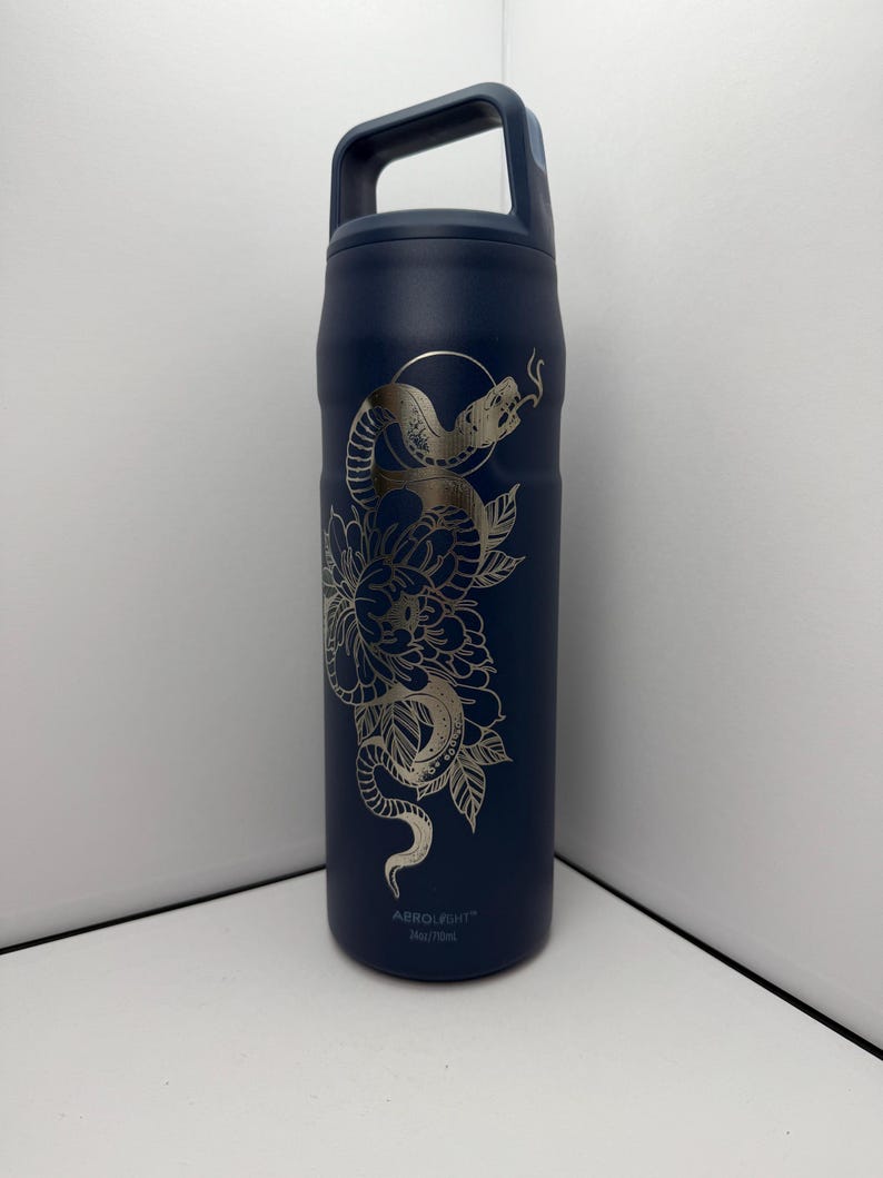 May include: A navy blue insulated water bottle with a silver floral and snake design. The bottle has a handle and the text "ABROLIGHT" and "740/750ml" printed on the bottom. The design features a snake coiled around a flower.