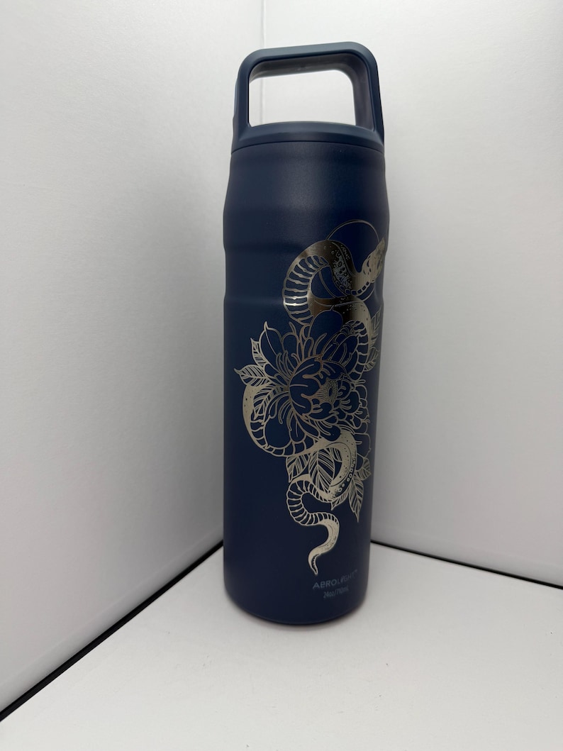 May include: Navy blue insulated water bottle with a silver floral and snake design. The bottle has a handle and a screw-on lid. The design wraps around the bottle, featuring a detailed flower and snake illustration.