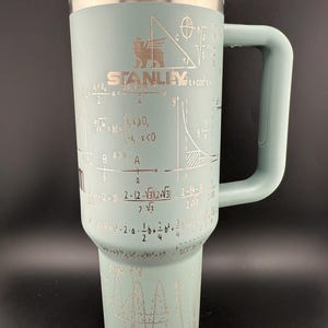 May include: A teal Stanley tumbler with a handle and clear lid. The tumbler features the Stanley logo and mathematical equations etched in silver. The design includes geometric shapes and formulas, creating a unique, scientific aesthetic.