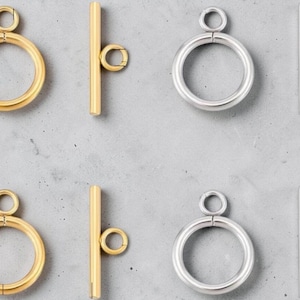 Può includere: Primo piano di chiusure a levetta dorate e argentate. Le chiusure presentano un anello circolare con un anello più piccolo attaccato e una barra dritta con un piccolo anello. Le chiusure sono esposte su una superficie grigia.