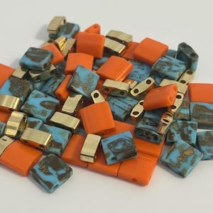 May include: A collection of beads in various shapes and colours. The beads include square orange beads, rectangular gold-coloured beads, and square beads with a blue and brown mottled pattern. The beads are scattered on a white surface.