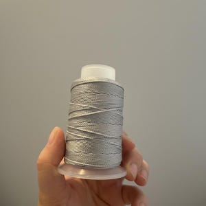Può includere: Una bobina di filo grigio con parte superiore bianca e base in plastica trasparente. Il filo è avvolto strettamente, mostrando una superficie strutturata. Ideale per cucito, ricamo o progetti artigianali.