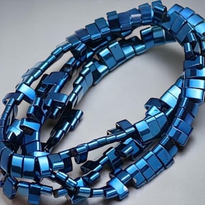 Op de afbeelding: Een ketting bestaande uit talrijke kleine, vierkante, metaalblauwe kralen. De kralen zijn aan elkaar geregen en vormen een flexibele ketting die buigt en oprolt. De ketting heeft een reflecterende, glanzende afwerking, tegen een neutrale achtergrond.