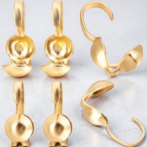 Puede incluir: Una colección de hallazgos de joyería dorados, que incluyen cierres y elementos decorativos. Los hallazgos tienen un acabado liso y pulido y están diseñados con formas curvas y redondeadas. Las piezas están dispuestas sobre una superficie gris claro.