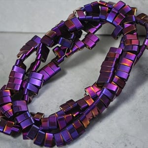 Puede incluir: Un collar de múltiples hilos compuesto por cuentas rectangulares e iridiscentes. Las cuentas cambian de color de púrpura a bronce, creando un efecto vibrante y llamativo. El collar está dispuesto en un patrón circular, mostrando su diseño.