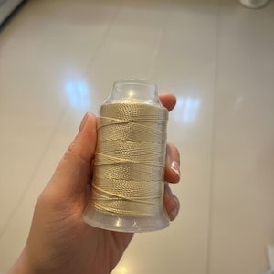 May include: A spool of beige thread, tightly wound around a clear plastic core. The thread has a slight sheen, suggesting a smooth texture. The spool is held in a hand, showcasing the thread's color and form. Ideal for sewing and crafting projects.