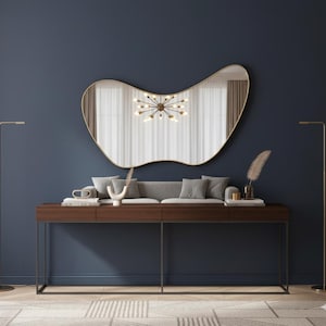 May include: A modern interior scene featuring a large, uniquely shaped gold-framed mirror above a dark wood console table. The table has a gray sofa, decorative items, and a pair of gold floor lamps. The wall is a deep blue.