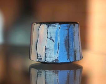 Puede incluir: Una taza de cerámica con un exterior texturizado y facetado con rayas verticales en azul claro, blanco y gris carbón oscuro. El recipiente tiene una forma cilíndrica ligeramente cónica con un borde oscuro y una base estrecha.