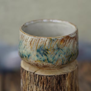 May include: A small, handcrafted ceramic bowl with a textured surface. The bowl features a blend of colours, including blue, green, and brown, with a cream-coloured interior. It sits atop a wooden tree stump, showcasing its rustic charm.