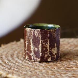May include: A cylindrical ceramic cup featuring a textured, dark brown exterior with vertical, distressed cream-coloured streaks. The interior of the cup is glazed in a smooth, deep green finish, contrasting with the rough, earthy outer surface.