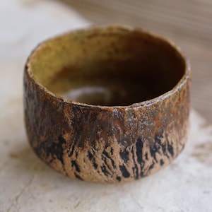 May include: A handcrafted ceramic bowl with a textured, earthy aesthetic. The bowl has a dark brown exterior with black markings and a lighter brown interior. The rim is rounded, and the bowl rests on a neutral-coloured surface.