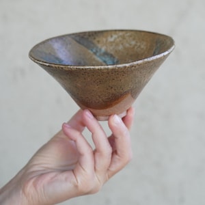 May include: A handmade ceramic bowl with a speckled brown glaze. The bowl has a wide, angled rim and a small, rounded base. The interior features streaks of blue and brown. The bowl is held in a hand.