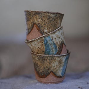 May include: Four handmade ceramic cups stacked together. The cups feature a speckled, earthy brown glaze with blue accents and a pinkish-brown base. The cups are stacked on top of each other.