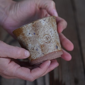 May include: A handmade ceramic cup with a speckled, textured surface. The cup has a light brown base and a lighter, speckled body with a stamped design. The cup is held in two hands.