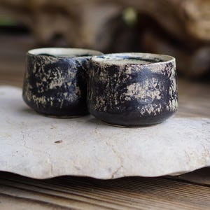 Puede incluir: Dos tazas de cer&aacute;mica peque&ntilde;as y redondas con un exterior negro oscuro y un acabado beige texturizado y desgastado. El interior de cada taza es liso y de color claro, contrastando con la superficie exterior oscura y rugosa.