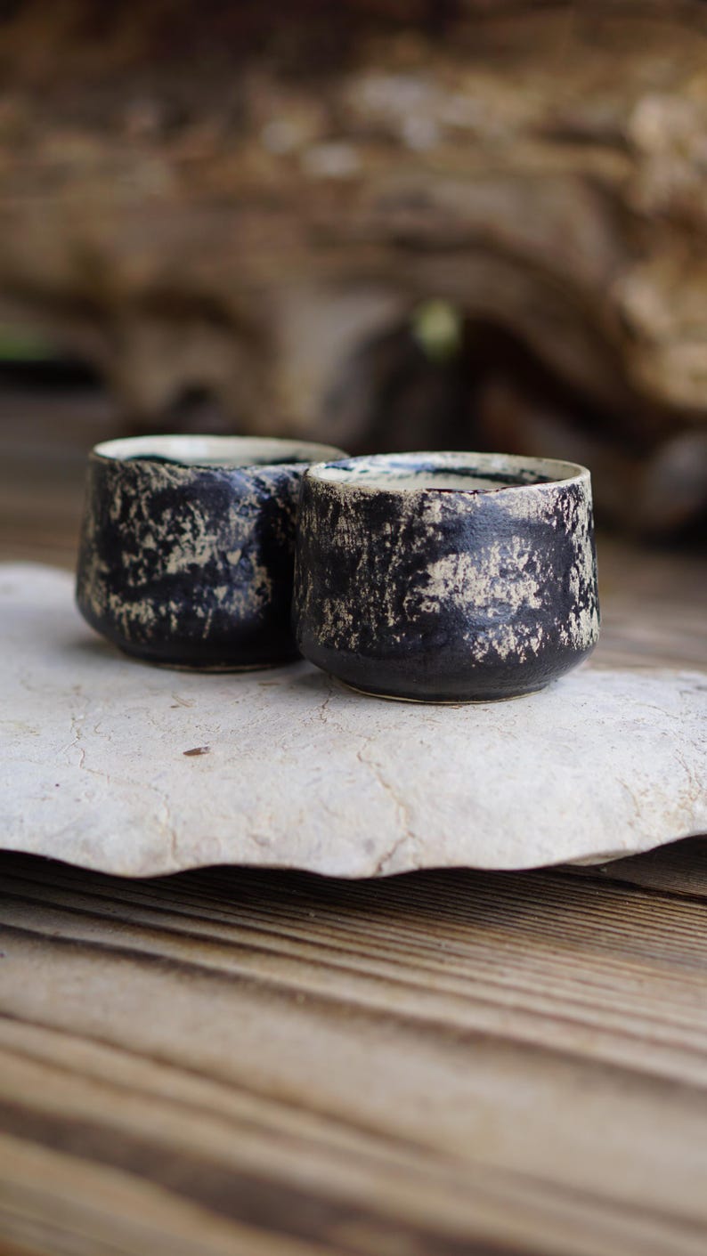 Puede incluir: Dos tazas de cer&aacute;mica peque&ntilde;as y redondas con un exterior negro oscuro y un acabado beige texturizado y desgastado. El interior de cada taza es liso y de color claro, contrastando con la superficie exterior oscura y rugosa.