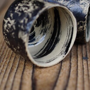 Puede incluir: Dos tazas de cer&aacute;mica con un exterior negro texturizado con motas de color crema y un interior liso decorado con pinceladas negras audaces y amplias sobre una base crema. Las tazas tienen una forma de borde irregular y org&aacute;nica.