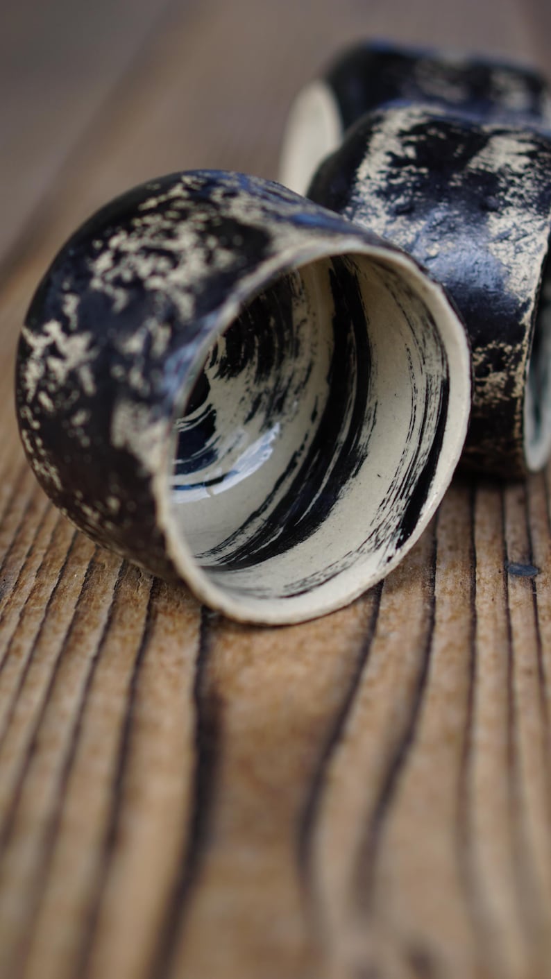 Puede incluir: Dos tazas de cer&aacute;mica con un exterior negro texturizado con motas de color crema y un interior liso decorado con pinceladas negras audaces y amplias sobre una base crema. Las tazas tienen una forma de borde irregular y org&aacute;nica.