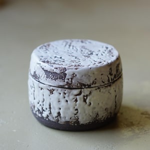 May include: A small, cylindrical ceramic jar with a lid, featuring a dark brown base clay partially covered in a thick, textured, white glaze that creates a rugged, uneven appearance across the exterior surface of both the body and the lid.