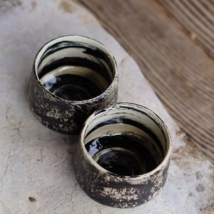 Puede incluir: Dos tazas peque&ntilde;as de cer&aacute;mica con forma redondeada. El exterior presenta un acabado texturizado y moteado en negro y blanco roto. El interior est&aacute; esmaltado con patrones de pinceladas horizontales en negro y crema en las paredes.