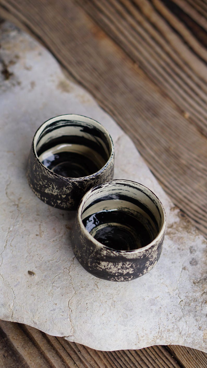 Puede incluir: Dos tazas peque&ntilde;as de cer&aacute;mica con forma redondeada. El exterior presenta un acabado texturizado y moteado en negro y blanco roto. El interior est&aacute; esmaltado con patrones de pinceladas horizontales en negro y crema en las paredes.