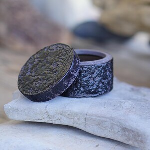 May include: A small, round, dark purple ceramic container with a matching lid. The exterior surface of both the lid and the base features a heavily textured, rugged, and uneven finish that resembles volcanic rock or rough, weathered stone.