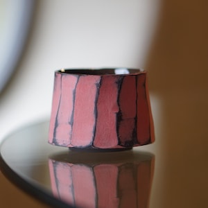 Può includere: Una piccola ciotola in ceramica fatta a mano con un design unico. La ciotola presenta un esterno strutturato rosa-rossastro con linee verticali scure. L'interno è scuro. La ciotola si riflette su una superficie di vetro.