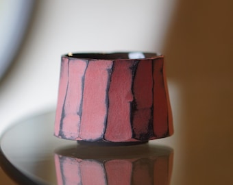Puede incluir: Una pequeña taza de cerámica cilíndrica con un exterior texturizado de color rosa coral mate que presenta rayas verticales hundidas de color carbón oscuro. El borde es ligeramente irregular y el interior tiene un acabado esmaltado oscuro.