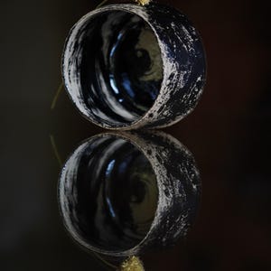 Puede incluir: Una taza de cer&aacute;mica con un acabado de esmalte jaspeado en blanco y negro, con un exterior rugoso y texturizado y un interior liso. Una peque&ntilde;a flor seca de color amarillo p&aacute;lido est&aacute; colocada en el borde de la taza, reflejada debajo.